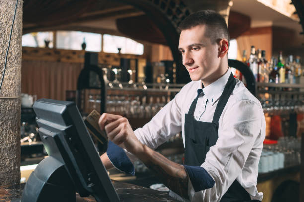small business, people and service concept - happy man or waiter in apron at counter with cashbox working at bar